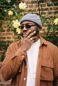 a man wearing a brown jacket and beanie is leaning against a brick wall